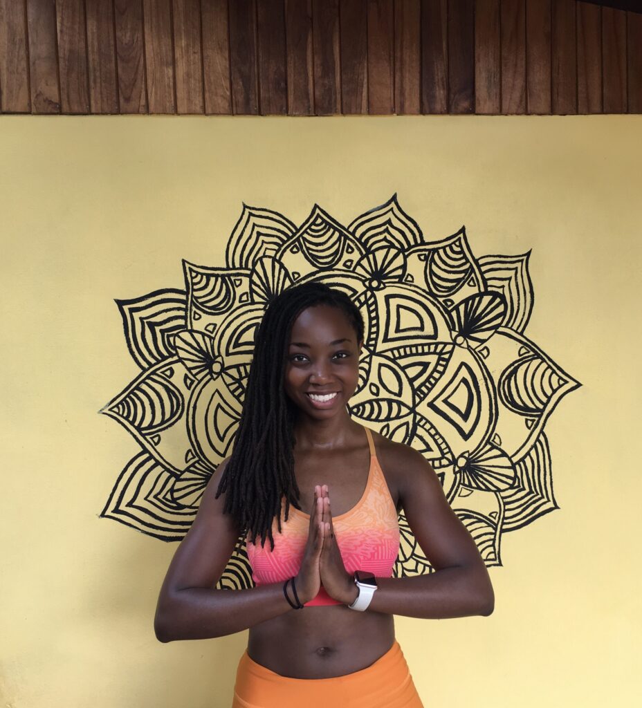 black yoga teacher Erica Rascon with namaste mudra in front of a mandala painting