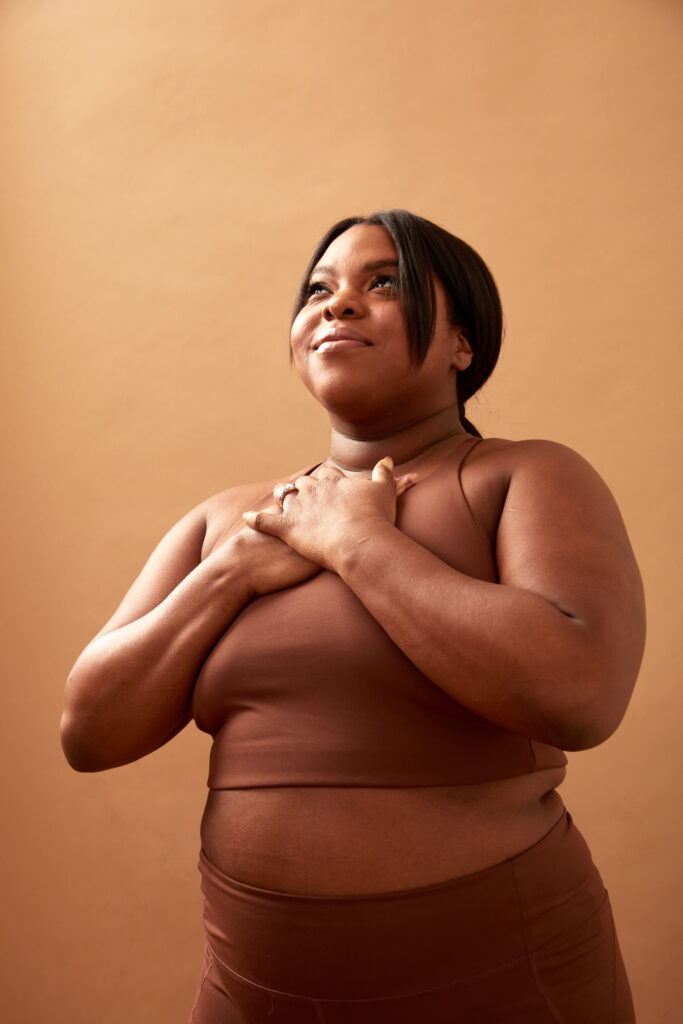 Black woman with full figure placing her hands over her chest. She gazes above her with a content, gentle smile. She stands before a rust-colored background.