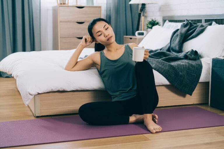 japanese female adult sitting in yoga outfit taking a break with hot drink by bedside. asian millennial woman closing her eyes and holding head by hand on bed is resting after workout.