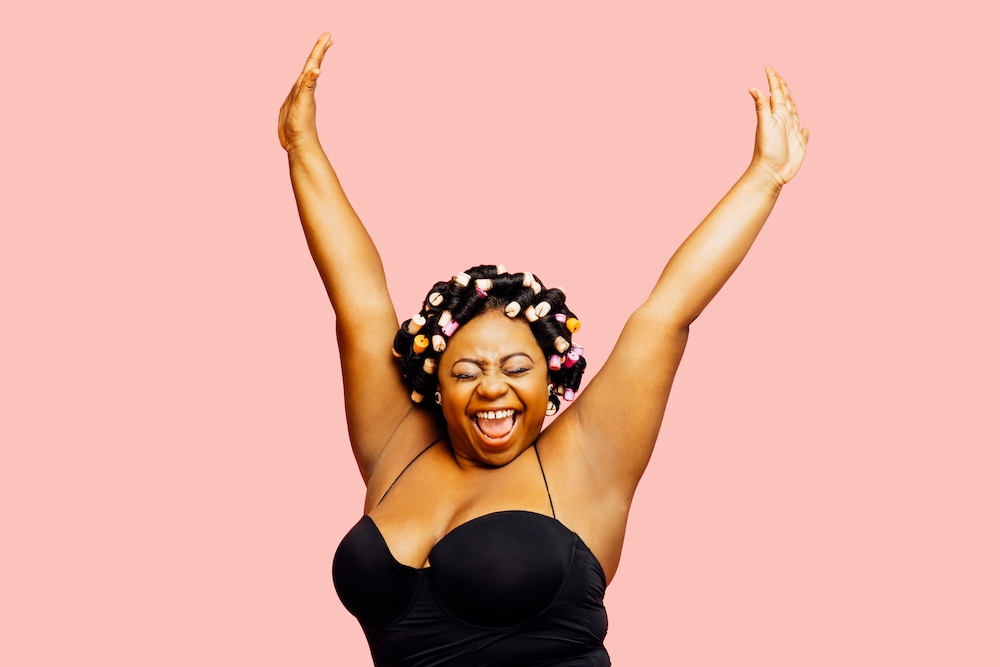 photo of full-figured black woman with rollers in her hair cheering over a solid, peach background
