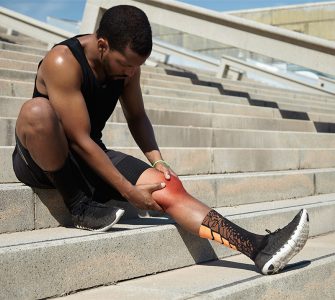 Portrait of dark-skinned athlete in black training outfit relaxing on concrete stairs after jogging exercises during his morning run, having pain in leg, touching his aching knee depicted in red color